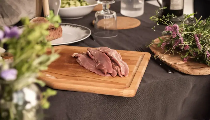 Aiguillettes de canard crues sur une planche à découper en bois, prêtes à être cuisinées. Des herbes fraîches et du pain complètent la scène.