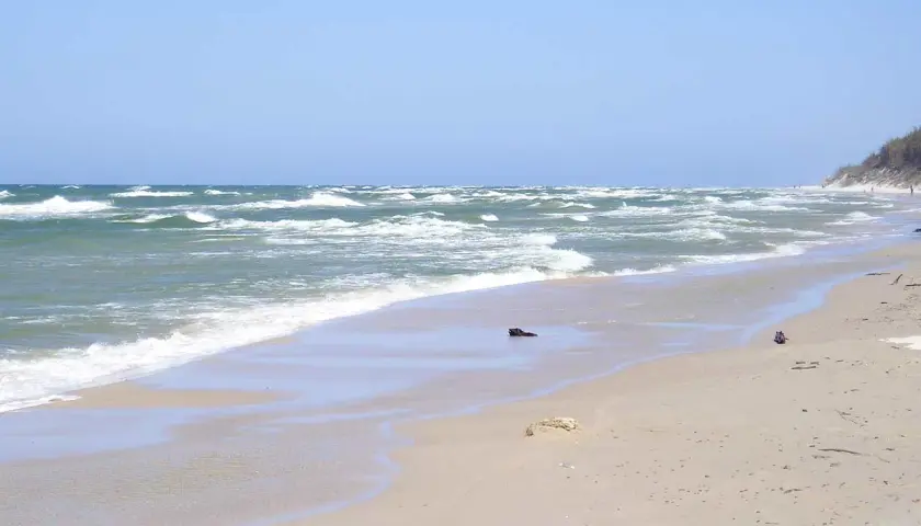 Plaża w Rowach, z falującym morzem i piaszczystym brzegiem. W oddali widać wydmy i nielicznych spacerowiczów.