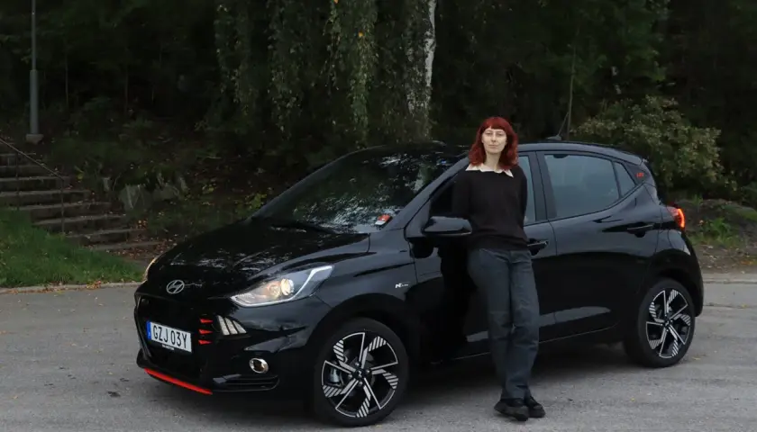 En kvinna med rött hår lutar sig mot en svart Hyundai i10. Bilen är redo för ett test.
