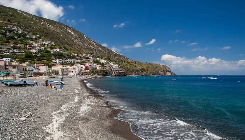 Vue d'une plage de galets sur l'île de Lipari, avec des maisons colorées accrochées à la colline et des bateaux dans la mer d'un bleu profond.
