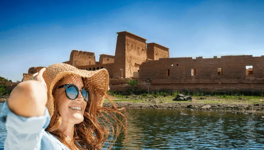 Mujer con sombrero y gafas de sol sonríe en un barco, con templos egipcios de fondo. ¡La mejor época para viajar a Egipto es ahora!