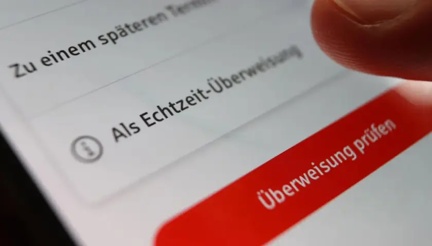 Finger tippt auf "Überweisung prüfen" auf einem Smartphone. Die Option "Als Echtzeit-Überweisung" ist sichtbar. Banken buchen oft sofort.