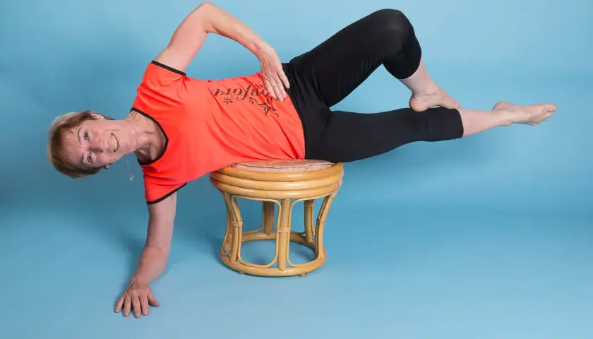 Ältere Frau in orangefarbenem Shirt und schwarzen Leggings macht eine seitliche Übung auf einem Hocker. Ihr linker Unterschenkel ist angewinkelt, der rechte gestreckt.