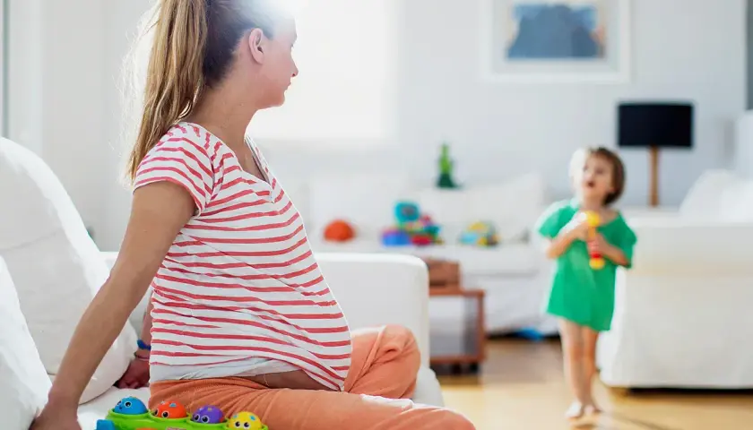 Schwangere Frau sitzt auf Sofa, Kind spielt. Frage: haushaltshilfe nach op wie viele stunden?