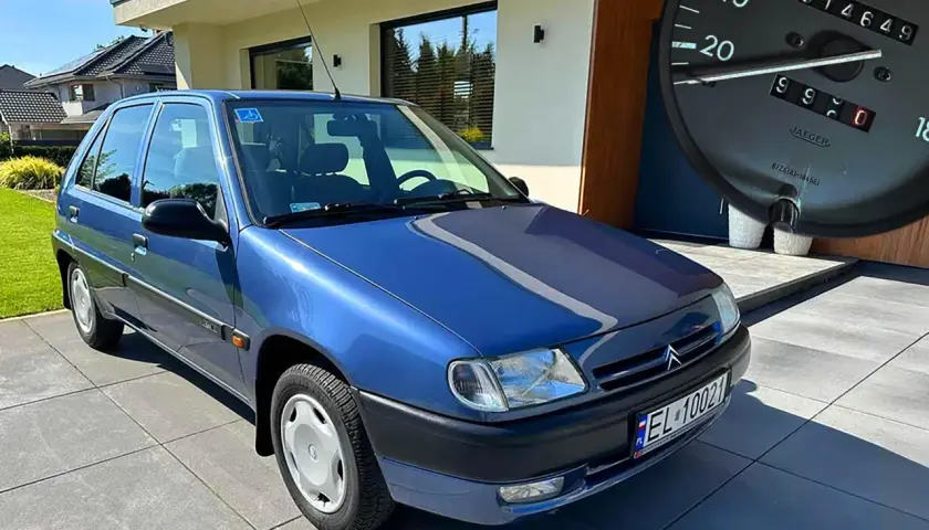 Niebieski Citroen Saxo z niskim przebiegiem, zaparkowany przed nowoczesnym domem. Na desce rozdzielczej widać licznik z przebiegiem 14649 km.