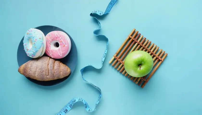 Dwa pączki i rogalik na talerzu obok zielonego jabłka i miarki krawieckiej. Wybór między słodyczami a zdrowiem, między kj a kcal.