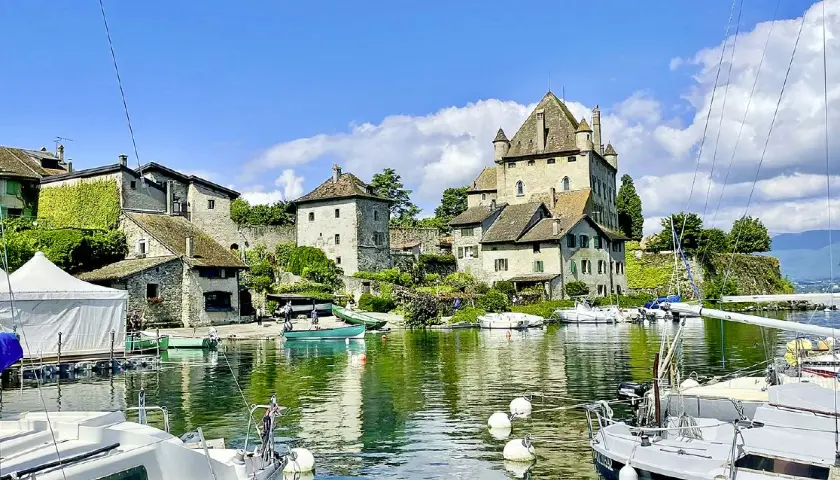 Vue d'une aire de camping-car au bord du lac Léman, avec un château médiéval, des bateaux et des maisons en pierre.