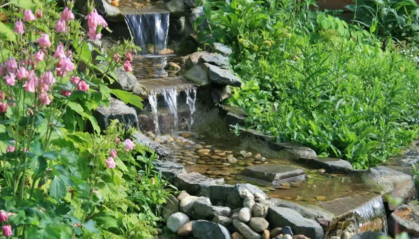 Ein kleiner, mehrstufiger Wasserfall im Garten, umgeben von üppigem Grün und rosa Blüten. Tolle wasserfall garten ideen für eine entspannende Oase.