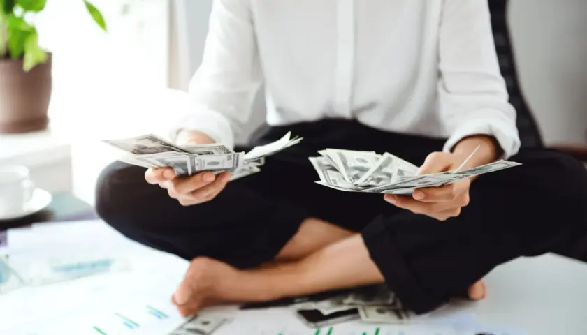 Osoba liczy banknoty dolarowe, przygotowując się do rozliczenia **umowy o dzieło podatek**. Na stole leżą wykresy i pieniądze.