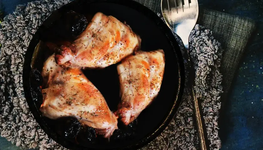 Trois morceaux de lapin aux pruneaux, assaisonnés et dorés, servis dans un plat noir avec une cuillère argentée.