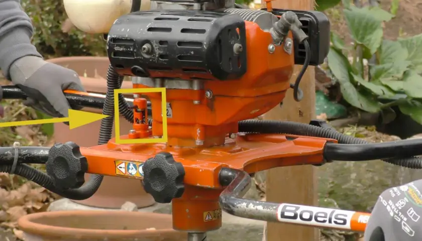 Ein orangefarbener Erdbohrer im Garten, bereit für den Einsatz. Ein gelber Pfeil hebt einen roten Hebel hervor.