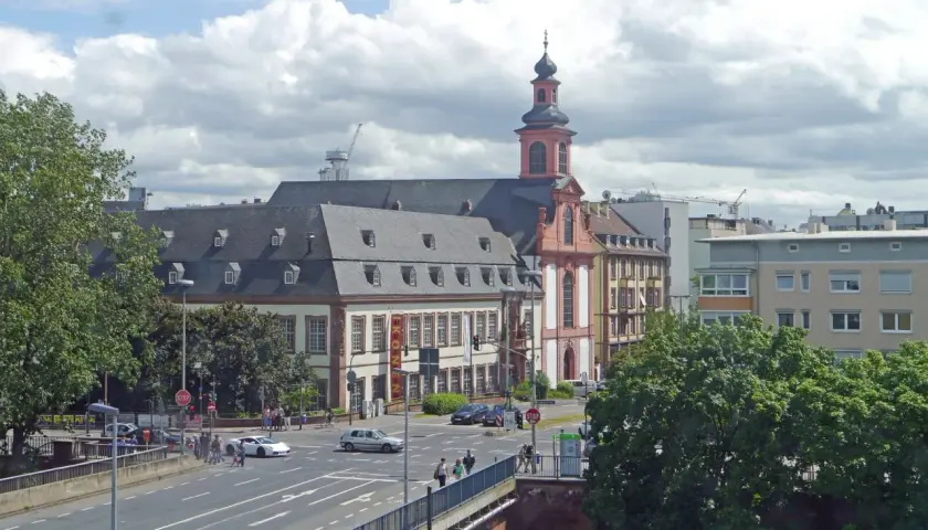 Barocke Kirche mit Turm und Mansarddach, Teil des Jakobsweg Hessen. Autos fahren auf einer Straße, Menschen überqueren sie.