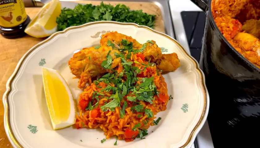 En tallrik med arroz con pollo, kyckling och ris med tomat, garnerad med persilja och citronklyfta. En gryta med samma rätt syns i bakgrunden.