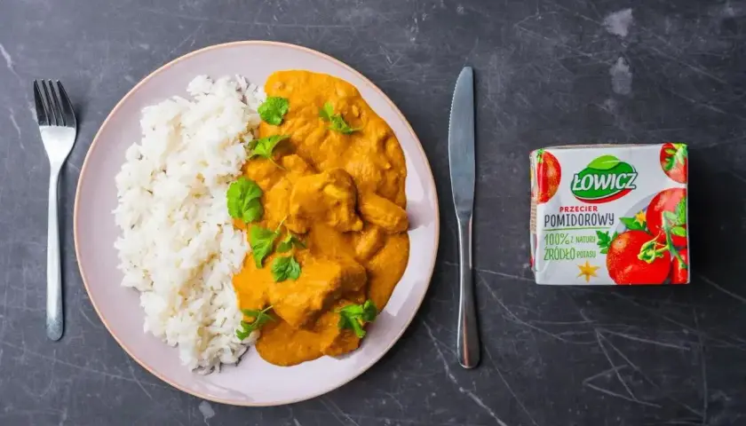 Pyszny butter chicken przepis na talerzu z ryżem, udekorowany świeżą kolendrą. Obok nóż, widelec i przecier pomidorowy Łowicz.