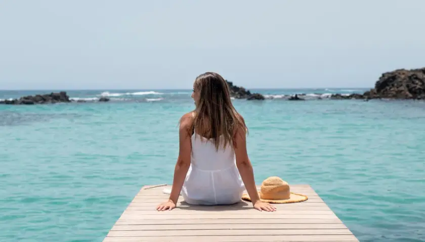 Mujer con vestido blanco sentada en un muelle de madera, disfrutando de la vista al mar turquesa. Su sombrero de paja descansa a su lado, perfecta para viajar sola.