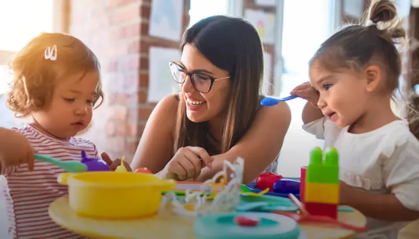 Eine lächelnde Frau mit Brille spielt mit zwei Kleinkindern. Sie lernt den erzieher berufsbegleitend und fördert spielerisch die Entwicklung der Kinder.