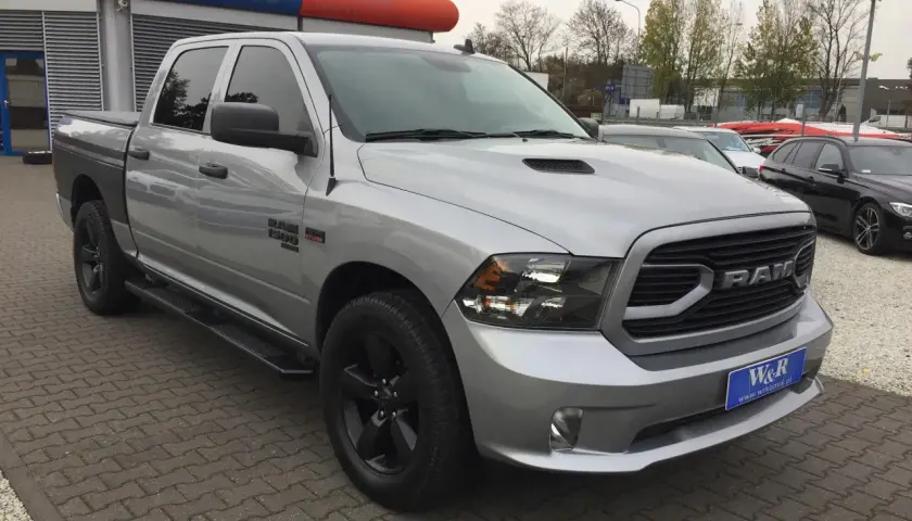 Srebrny Dodge Ram 1500 5.7 Hemi z czarnymi felgami i przyciemnianymi szybami stoi na parkingu przed budynkiem z niebiesko-pomarańczowym banerem.