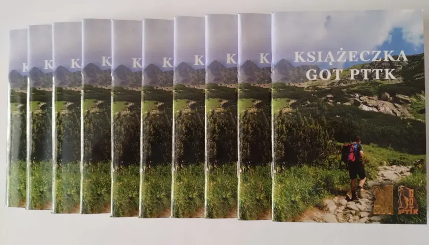 Rząd książeczek GOT PTTK z widokiem na góry i turystę na szlaku.