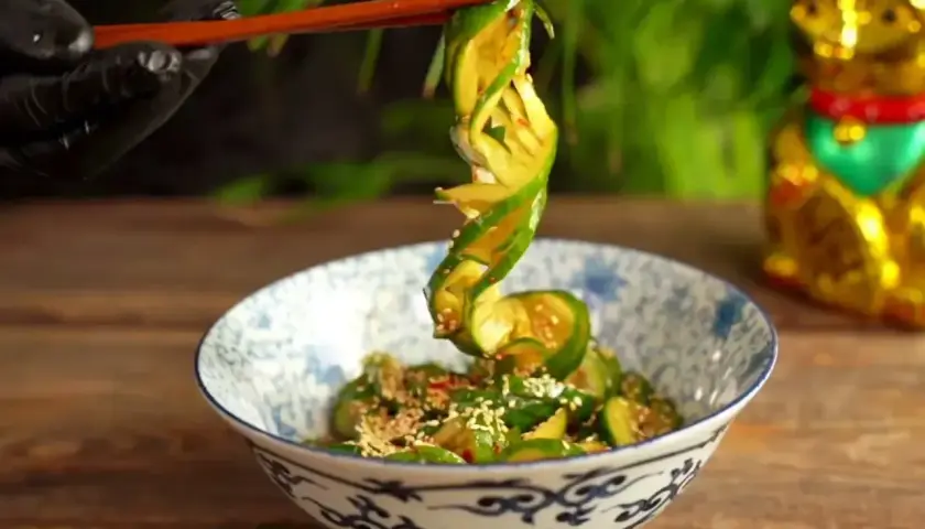 Chopsticks podnoszą zwinięty plasterek ogórka z miski pełnej tajskiej sałatki z ogórka, posypanej sezamem.