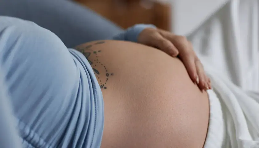 Dłoń delikatnie spoczywa na **twardym brzuchu w ciąży**, ozdobionym subtelnym tatuażem.