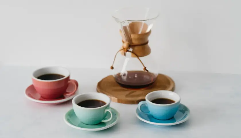Chemex vs drip: trzy kolorowe filiżanki z kawą i szklany dzbanek Chemex na drewnianej podstawce.
