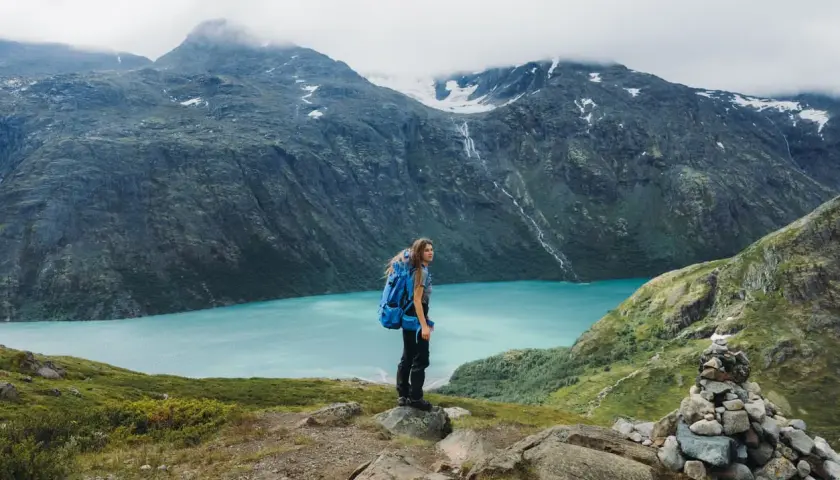 A hiker with a blue backpack stands on a rocky outcrop, overlooking a stunning turquoise lake in Jotunheimen National Park. The dramatic mountains behind are shrouded in mist.
