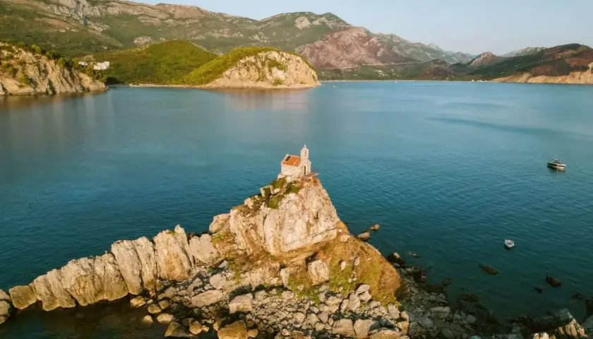 A tiny church perches on a rocky islet, a picturesque stop on a Montenegro itinerary. Mountains rise behind the tranquil blue bay.
