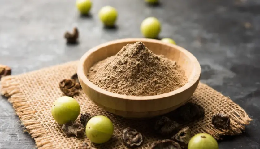 Polvere di triphala in una ciotola di legno, con accanto frutti di amla e parti essiccate. Il triphala a cosa serve? Aiuta la digestione e il benessere generale.
