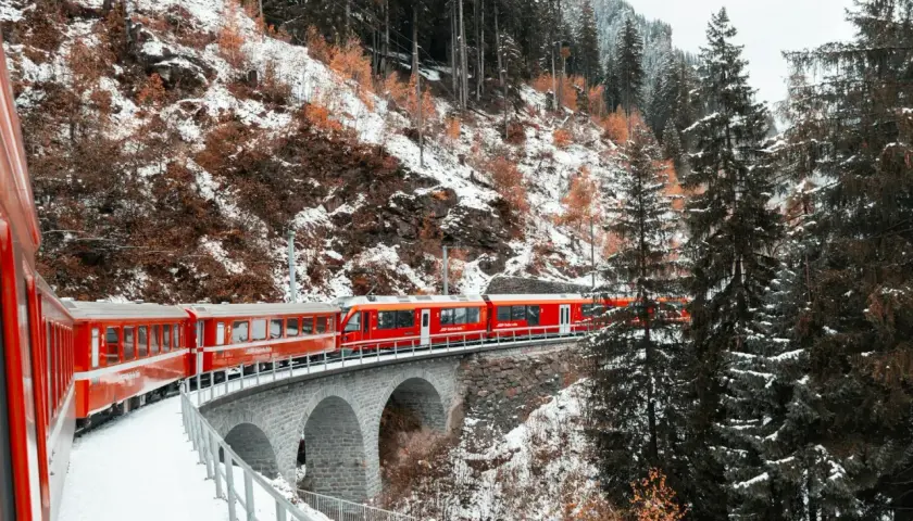 Czerwony pociąg Glacier Express przejeżdża przez kamienny most w zimowym, zaśnieżonym krajobrazie górskim.