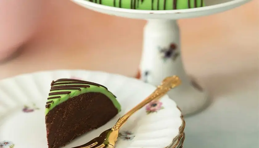 En bit av en himmelsk hallontårta med grön marsipan och chokladdrizzle. En gyllene gaffel ligger på en tallrik med blommor.