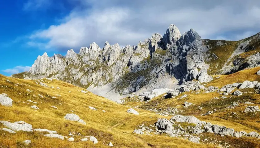 A winding path leads through golden grass and rocks towards the jagged peaks of Bobotov Kuk, a stunning hike under a dramatic sky.