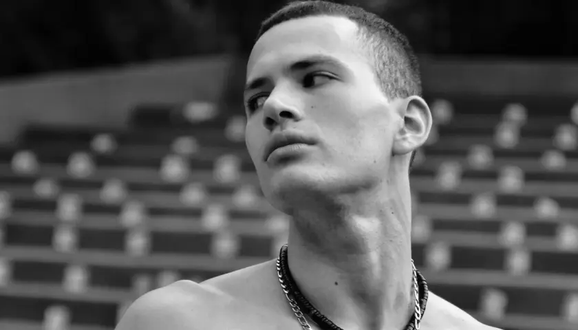 Joven con buzz cut largo, collar plateado y mirada perdida al horizonte.