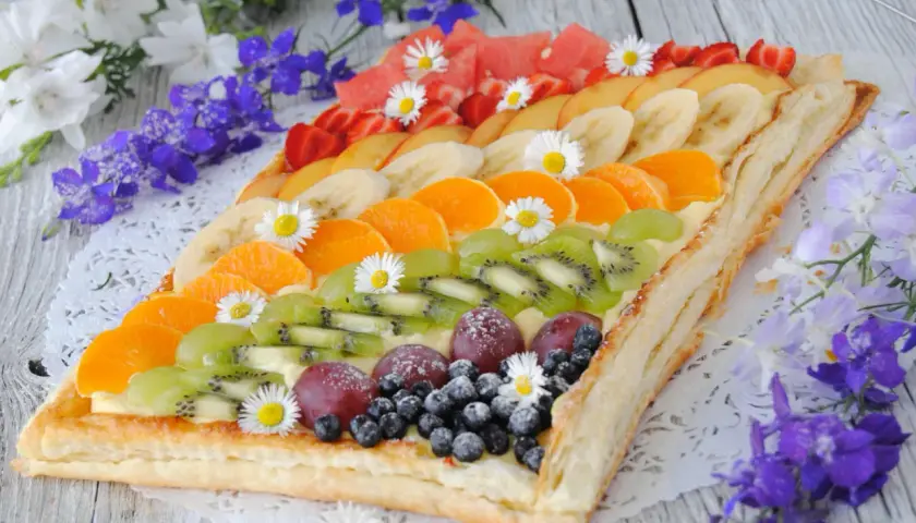 Kolorowa tarta z owocami: kiwi, mandarynki, banany, truskawki, winogrona i borówki, ozdobiona stokrotkami.