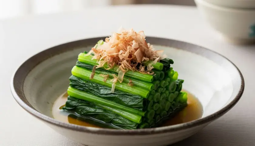 A neat stack of blanched spinach, known as spinach ohitashi, is served in a shallow bowl with a light broth and topped with delicate bonito flakes.