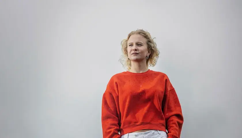 Sigrid Johnson, med sitt blonda, lockiga hår, bär en orange tröja och tittar drömmande bort.