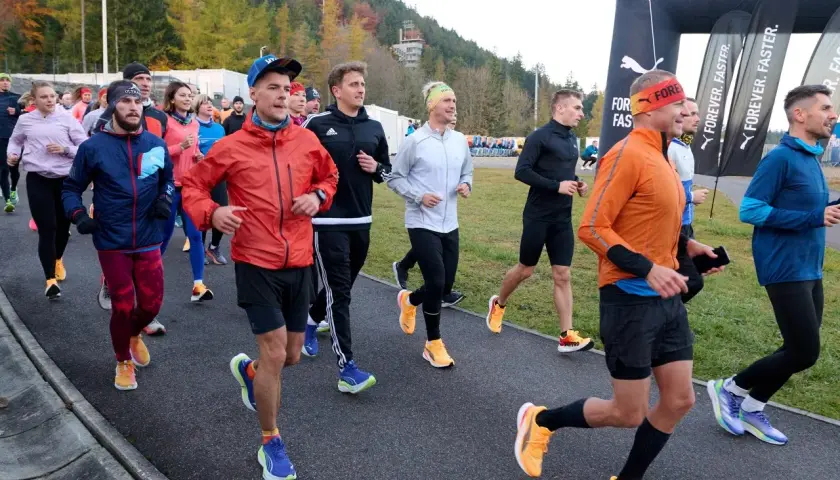 Grupa biegaczy w kolorowych strojach, jak z filmu o bieganiu, startuje na trasę. W tle las i bramka mety Puma.