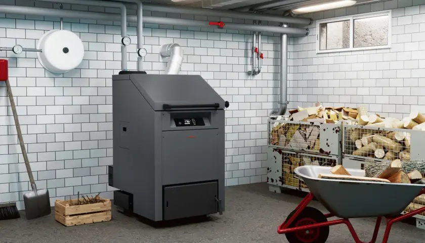 Kotłownia z nowoczesnym piecem na biopaliwo, obok składowane drewno opałowe w metalowych koszach i taczka z polanami.