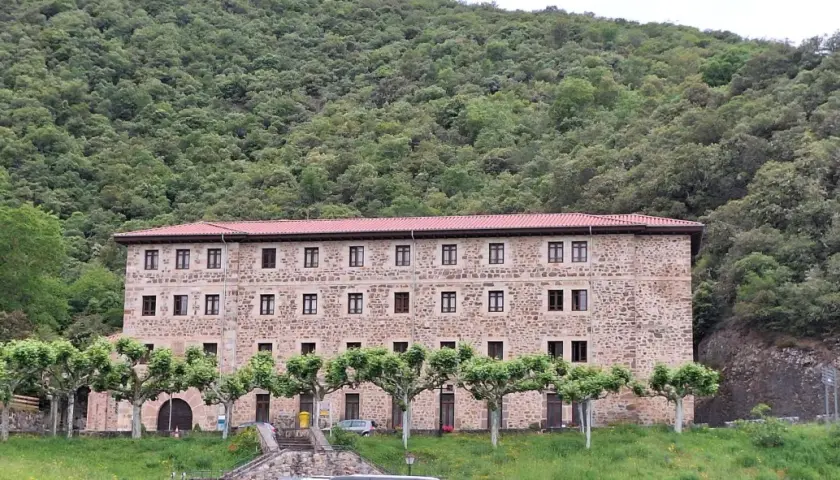 Edificio de piedra con tejado rojo, rodeado de árboles y un monte frondoso. Es el monasterio de Santo Toribio de Liébana.