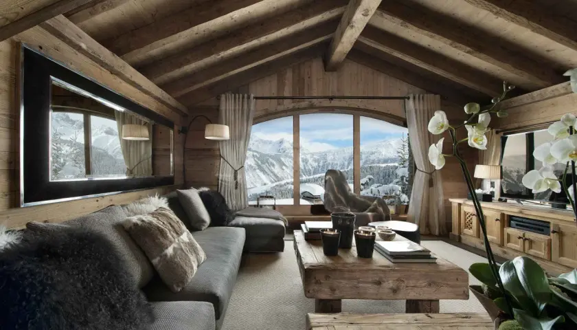 Gemütliches Wohnzimmer mit rustikaler Holzwand und Decke, einem großen Fenster mit Bergblick, einem Sofa mit Fellkissen und einem Holztisch. Luxus Chalet Inneneinrichtung.