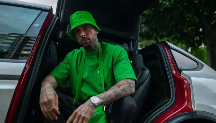 Malik Montana w zielonym kapeluszu i bluzie siedzi w otwartym samochodzie. Jego tatuaże i zegarek przyciągają wzrok, a jego wzrost jest imponujący.