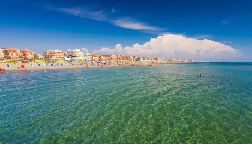 Słoneczny dzień nad morzem w Rzymie. Tłumy plażowiczów, kolorowe parasole i budynki wzdłuż wybrzeża.