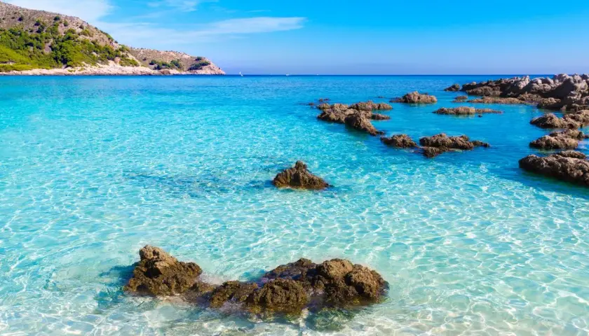 Eau turquoise cristalline, rochers émergeant de la mer, colline verdoyante en arrière-plan. Un rêve de toscane plage.