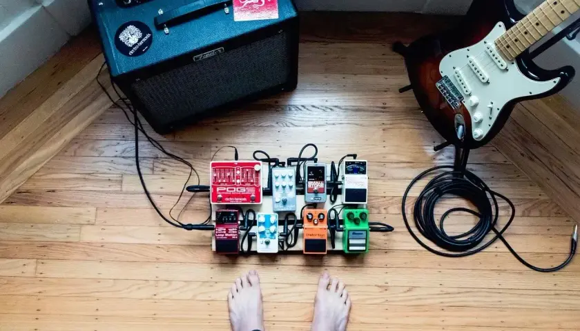 Stojący na drewnianej podłodze pedalboard z efektami gitarowymi, gotowy do gry. W tle wzmacniacz i gitara.