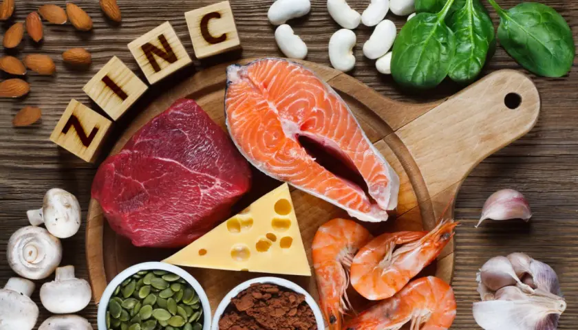 Zink-reiche Lebensmittel wie Rindfleisch, Lachs, Garnelen, Kürbiskerne, Käse, Spinat, Mandeln, Pilze und Knoblauch sind auf einem Holztisch arrangiert.