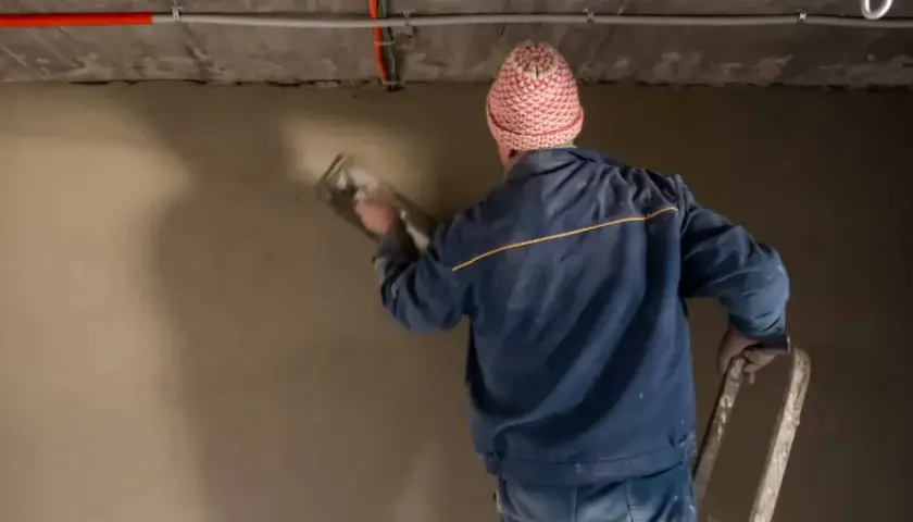 Mann mit Mütze verputzt eine Wand im Keller. Der **Sanierputz** im **Keller** wird sorgfältig aufgetragen.