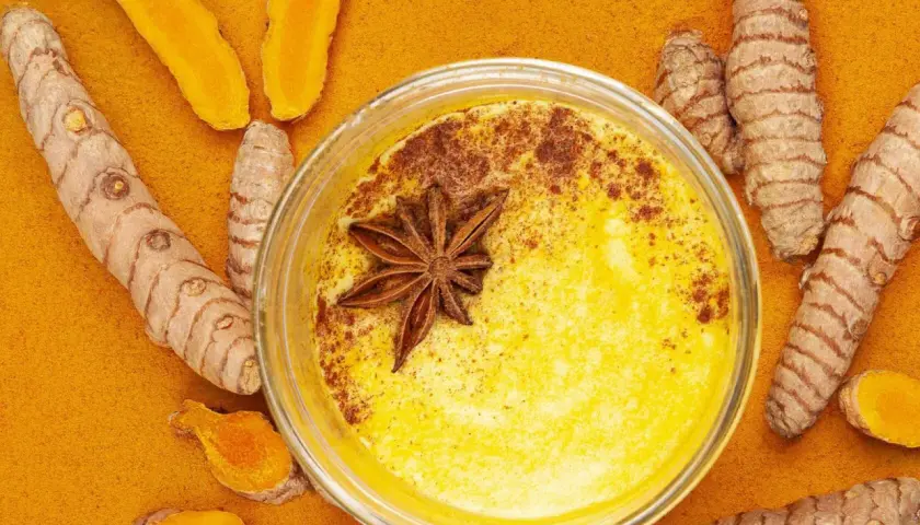 Goldene Milch mit Kurkuma und Honig, verziert mit Sternanis. Umgeben von frischen Kurkumawurzeln und Pulver.
