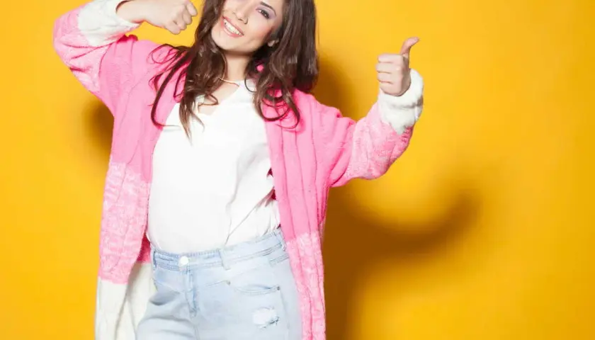 Une femme souriante, petite et ronde, montre comment s'habiller avec un gilet rose et un jean. Elle fait un signe de pouce levé.