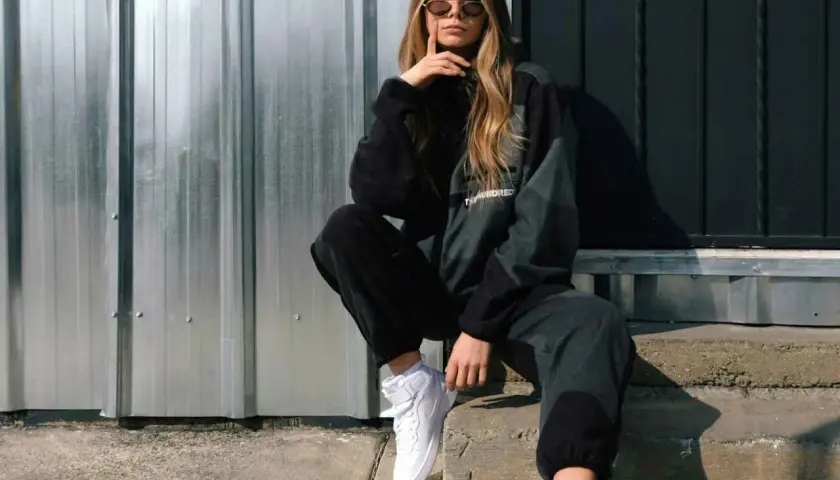 Chica con estilos de ropa mujer modernos, posa con sudadera y pantalones deportivos, zapatillas blancas y gafas de sol.