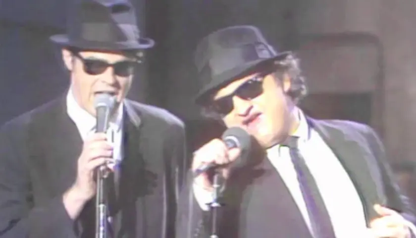 Dan Aykroyd giovane e John Belushi sul palco, entrambi con cappello e occhiali da sole, cantano al microfono.