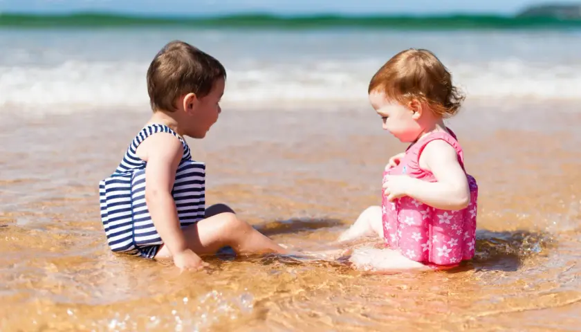 Dwoje dzieci w kostiumach kąpielowych z kamizelkami pływającymi siedzi na plaży.